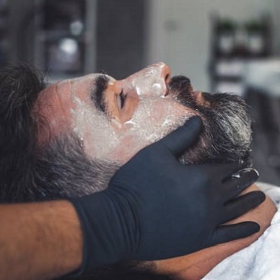 Hairdresser wearing latex gloves giving a facial to a handsome bearded man.